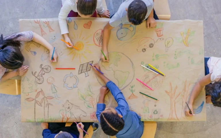 Crianças desenhando temas ambientais como reciclagem, energia solar e natureza em um papel grande. Atividade educativa que promove a conscientização ambiental.